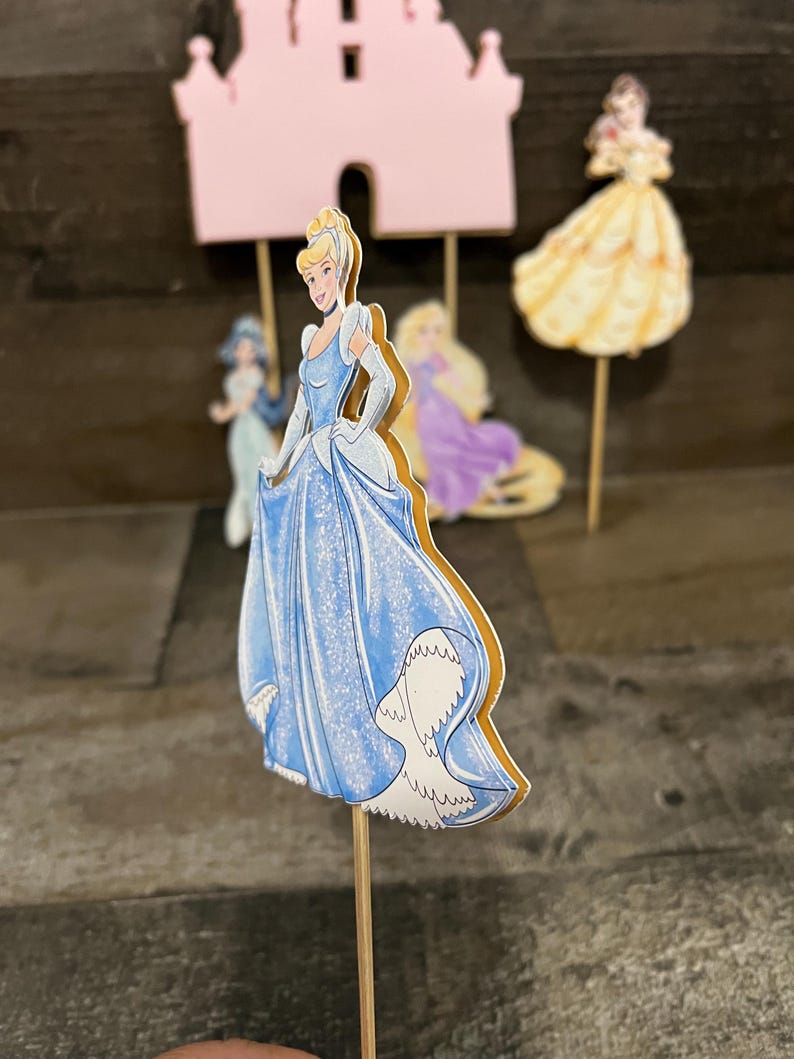 May include: A blue and white Cinderella cake topper with a sparkly blue dress and a golden trim. The topper is on a wooden stick.