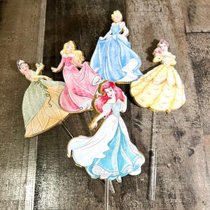May include: Five Disney princess cake toppers, each featuring a different princess in a colorful gown. The princesses are Cinderella, Belle, Ariel, Aurora, and Tiana. The toppers are made of cardboard and have a gold glitter finish.
