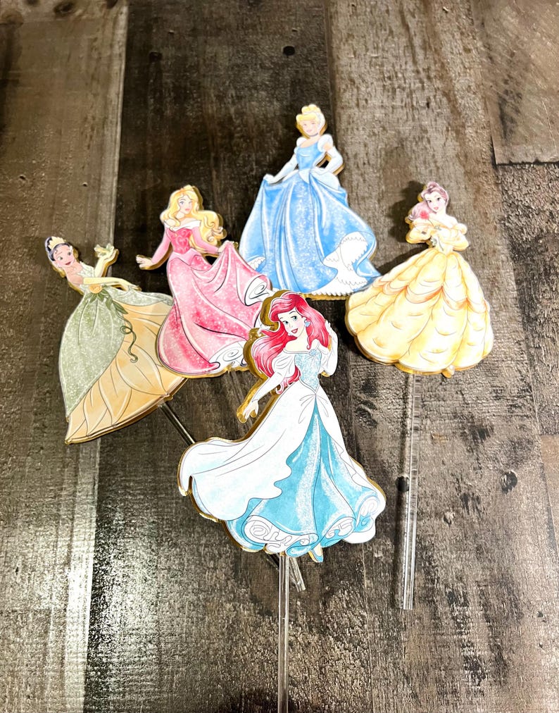May include: Five Disney princess cake toppers, each featuring a different princess in a colorful gown. The princesses are Cinderella, Belle, Ariel, Aurora, and Tiana. The toppers are made of cardboard and have a gold glitter finish.