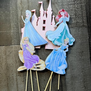 May include: Four princess cake toppers featuring Cinderella, Ariel, Rapunzel, and Elsa. Each topper is on a wooden stick and features a pink castle cutout.