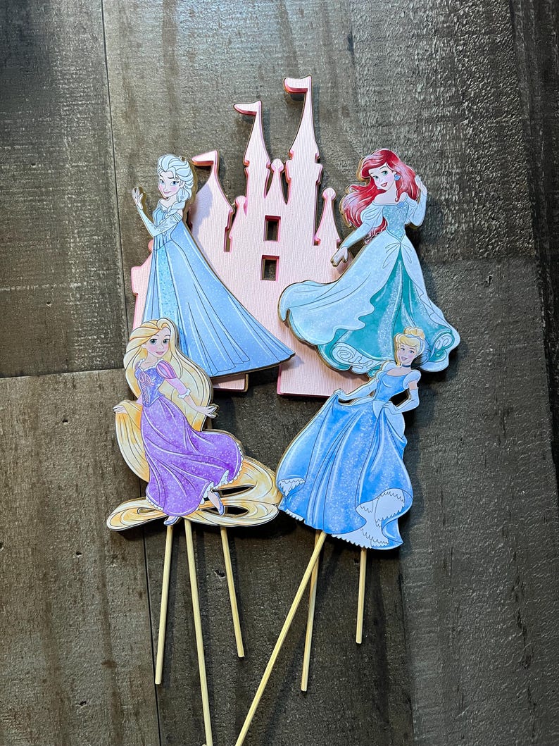 May include: Four princess cake toppers featuring Cinderella, Ariel, Rapunzel, and Elsa. Each topper is on a wooden stick and features a pink castle cutout.