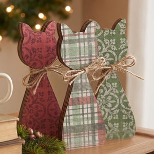 May include: Three cat-shaped wooden ornaments standing upright. Each figure features a distinct pattern: one in deep red with floral motifs, one in a green and red plaid, and one in green with white scrollwork. Each cat is tied with a jute twine bow.