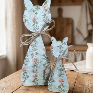 May include: Two cat-shaped wooden ornaments featuring a light blue background with a repeating pattern of pink roses and green vines. Each figure is adorned with a natural jute twine bow tied around the neck, standing upright on a flat base.