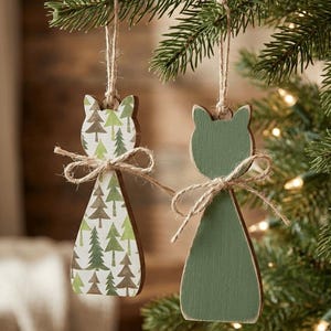 May include: Two cat-shaped wooden ornaments hanging from twine loops. One ornament features a green and brown pine tree pattern, while the other is solid sage green. Both ornaments are decorated with a small, tied twine bow at the neck area.