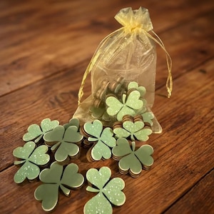 May include: A collection of green shamrock-shaped decorations with white speckles, scattered on a wooden surface. A small, sheer, gold-colored bag is filled with similar shamrock shapes. The shamrocks are a light green color.