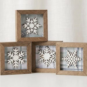 May include: Four square wooden frames arranged in a pyramid stack. Each frame contains a white snowflake cutout centred over a background featuring grey and white plaid or botanical patterns. The wood has a natural medium brown finish.