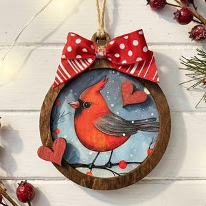 May include: A round wooden ornament featuring a red cardinal bird perched on a branch with red berries. The bird is set against a blue background with falling snow. A red and white polka dot bow and a twine loop are at the top. Two wooden hearts are attached to the ornament.