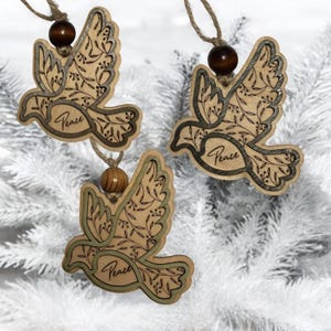 May include: Three wooden dove ornaments with the word "Peace" inscribed. The doves are light brown with a darker outline and a floral design. Each ornament has a wooden bead and twine for hanging.
