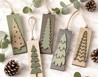 May include: Five rectangular wooden ornaments with jute twine loops, each featuring a unique cutout or layered tree design in shades of green, brown, and natural wood. The ornaments display varied tree shapes including geometric, scalloped, and textured silhouettes.