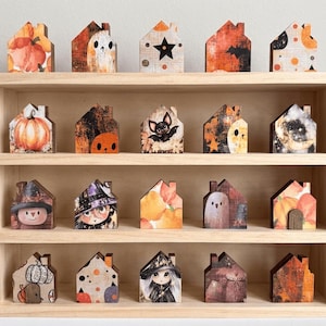 May include: A light-colored wooden shelf with four tiers holding twenty small, house-shaped wooden blocks. Each block features distinct Halloween-themed illustrations, including pumpkins, ghosts, bats, stars, moons, and characters in witch hats.