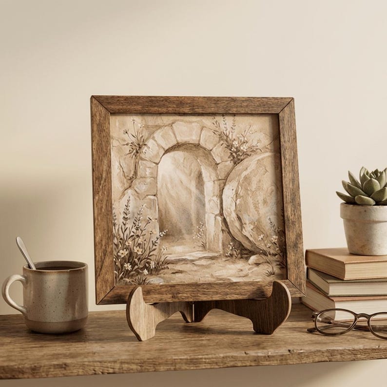 May include: A framed sepia-toned artwork depicting an arched stone doorway and a large stone. The artwork is displayed on a wooden easel. A ceramic mug with a spoon, a succulent in a pot, books, and glasses are also on the wooden shelf.