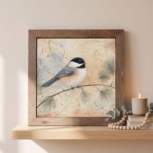 May include: A square framed wall art piece featuring a black-capped chickadee perched on a pine branch. The bird has a black head, white cheeks, and a soft tan and gray body. The background is a textured cream color with faint swirling lines.