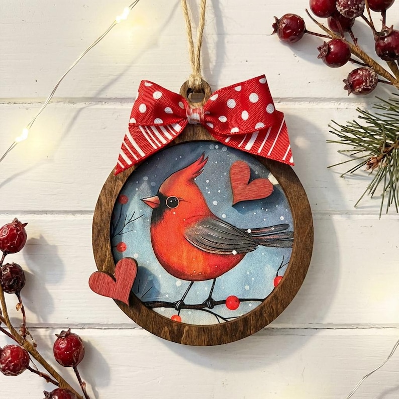 May include: A round wooden ornament featuring a red cardinal bird perched on a branch with red berries. The bird is set against a blue background with falling snow. A red and white polka dot bow and a twine loop are at the top. Two wooden hearts are attached to the ornament.