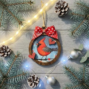 May include: A wooden Christmas ornament featuring a red cardinal bird illustration. The ornament has a red and white polka dot bow and two wooden heart accents. The background is a winter scene with snow and berries. The ornament is surrounded by pine branches and fairy lights.