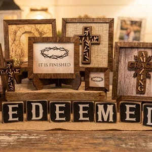 May include: A collection of framed wooden art pieces and decorative blocks. The frames feature crosses, botanical prints, and the words "He is Risen" and "It is Finished." The word "REDEEMED" is spelled out in individual blocks.