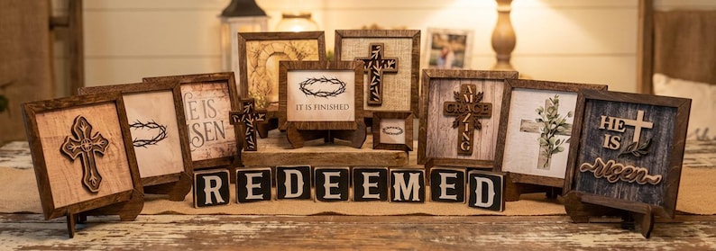 May include: A collection of framed wooden art pieces and decorative blocks. The frames feature crosses, botanical prints, and the words "He is Risen" and "It is Finished." The word "REDEEMED" is spelled out in individual blocks.