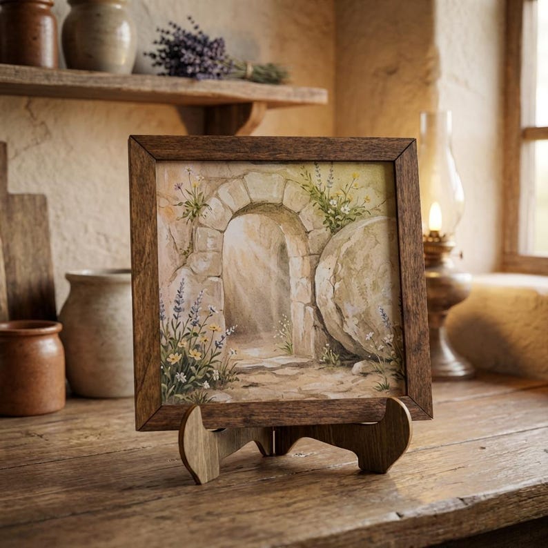 May include: A framed painting depicting an open stone tomb with a large stone rolled away. The artwork features an arched doorway, wildflowers, and a large stone. The frame and easel are made of wood.