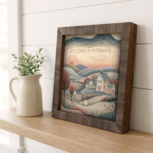May include: A square framed print featuring a landscape illustration of a house and rolling hills at sunrise with the text "JOY COMES IN THE MORNING" at the top. The frame is made of dark brown wood with a visible grain, surrounding the colorful artwork.