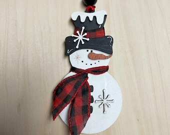May include: A wooden snowman ornament with a black top hat, a red and black plaid scarf, and a carrot nose. The snowman has a snowflake design and is hanging from a red ribbon. The ornament is approximately 4 inches tall.