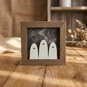 May include: A square wooden frame containing a dark, textured charcoal background with three white, rounded ghost-shaped cutouts. Each ghost figure features two small, black, oval-shaped eyes positioned near the top of the cutout.