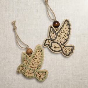 May include: Two wooden dove ornaments with a natural wood grain finish. Each dove has a dark outline and is filled with a floral design. The word "Peace" is written on each dove. The ornaments hang from twine with a wooden bead.