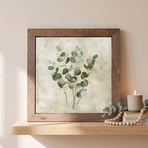 May include: A square wall art piece featuring a dark brown wooden frame surrounding a print of green eucalyptus branches against a mottled beige background. The botanical illustration displays multiple stems with rounded leaves in various shades of green.