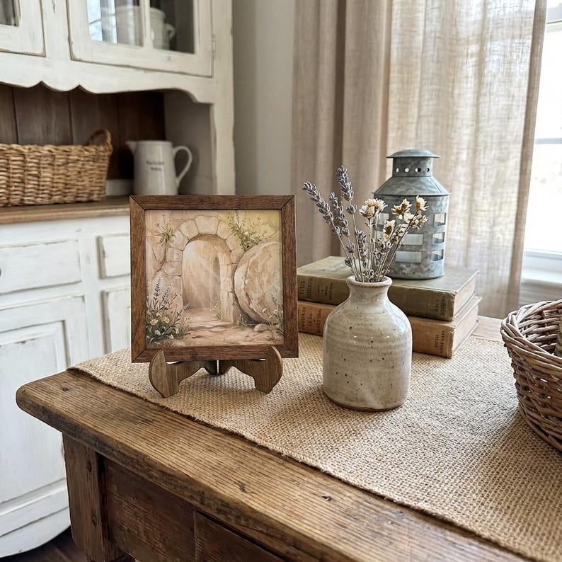 May include: A framed watercolor painting of an arched doorway with a stone exterior, displayed on a wooden easel. The painting is set on a rustic wooden table with a burlap runner. A small ceramic vase holds dried flowers, and a metal lantern sits nearby.