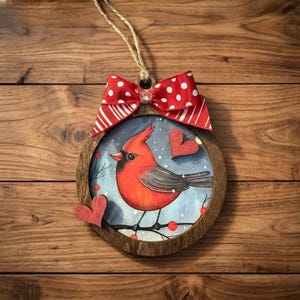 May include: Wooden Christmas ornament featuring a red cardinal bird illustration inside a round frame. The ornament has a red and white polka dot bow and a twine hanger. Two wooden hearts are attached to the frame.