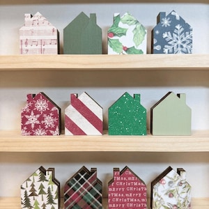 May include: Twelve small wooden house-shaped ornaments arranged on three shelves. Designs include musical notes, solid green, holly leaves, white snowflakes on blue, red snowflakes, red and white stripes, green speckles, light green, pine trees, plaid, text reading Merry Christmas, and winter berries.