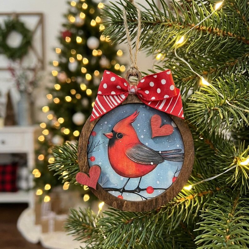May include: A round wooden Christmas ornament featuring a red cardinal bird illustration. The ornament has a red and white polka dot bow and two red heart accents. The ornament is hanging on a Christmas tree.