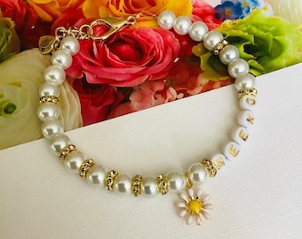 Puede incluir: Un brazalete de perlas blancas con un cierre dorado y un pequeño dije de margarita blanca. El brazalete tiene cuentas blancas con letras doradas que deletrean "EMMA."