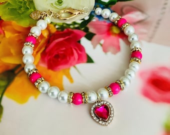 May include: A white pearl bracelet with pink beads and a gold heart charm with a red rhinestone. The bracelet is on a white surface with pink flowers in the background.