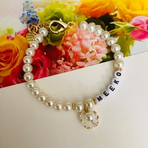 Puede incluir: Un brazalete de perlas blancas con un cierre dorado y un dije dorado en forma de concha. El brazalete tiene cuentas de letras blancas que deletrean el nombre "MEEKO".