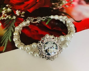 May include: A silver bracelet featuring a large, clear crystal set within a decorative silver frame. The bracelet is made of white pearls and has a silver clasp.