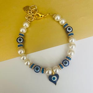 May include: A beaded bracelet featuring white faux pearls, blue and white evil eye beads, and small gold-toned spacers. The bracelet includes a heart-shaped evil eye charm and a gold-toned lobster clasp with a small heart-shaped tag attached.