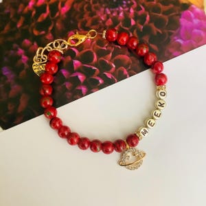 May include: A red beaded bracelet with gold accents and a charm. The bracelet features the name "MEEKO" in gold letter beads and a gold charm with a planet design. The bracelet is made with red beads and a gold clasp.