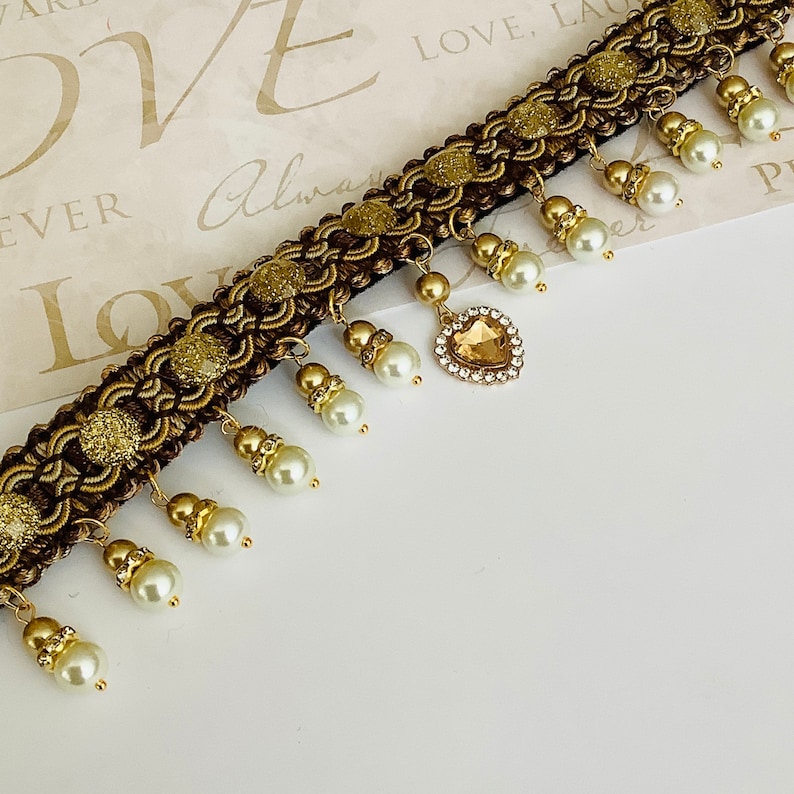 May include: A gold and brown beaded necklace with a heart-shaped pendant featuring a clear rhinestone. The necklace has a fringe of white pearls.