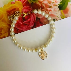 May include: A white pearl bracelet with a gold charm featuring the letter "B". The bracelet is accented with small gold beads and is displayed on a white surface.