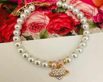 May include: A white pearl bracelet with a gold heart charm adorned with sparkling rhinestones. The charm features a small gold ring around it, resembling a planet.