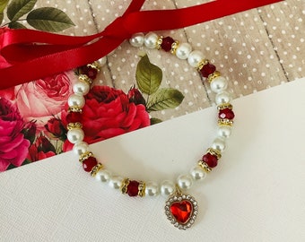 Puede incluir: Un collar de perlas blancas y rojas con un dije de corazón dorado y un cristal rojo en el centro. El collar está sobre un fondo blanco.