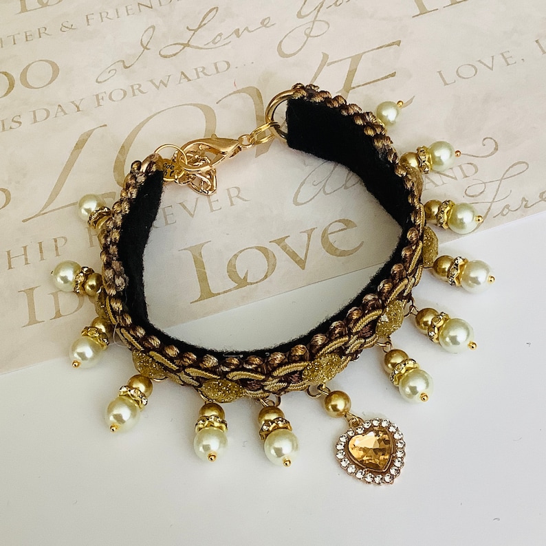 May include: A gold and black bracelet with a heart charm and pearl accents. The bracelet has a black velvet band and gold trim. The heart charm has a clear rhinestone center.