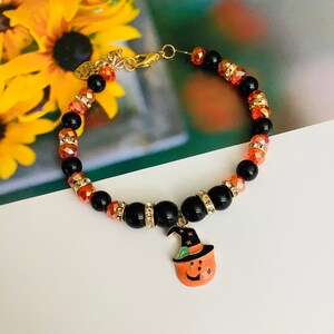 May include: A black and orange beaded bracelet with a pumpkin charm. The pumpkin charm has a green hat and a smiling face.
