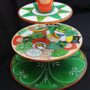 Può includere: Un porta torte in legno a tre livelli decorato con trifogli, arcobaleni e altre immagini del giorno di San Patrizio. Il livello superiore presenta una statuetta di folletto verde con la barba rossa.