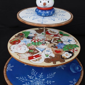 May include: A three-tiered wooden serving stand decorated with a winter theme. The top tier features a snowman with a black hat and a blue scarf. The middle tier is decorated with gingerbread cookies, candy canes, and other winter-themed designs. The bottom tier is painted blue with a white snowflake design.