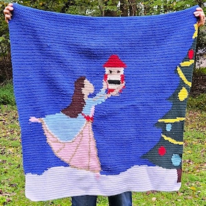 May include: A blue crocheted blanket featuring a ballerina holding a nutcracker, with a Christmas tree on the right. The ballerina wears a pink and blue dress. The blanket has a white border, and the background is blue.