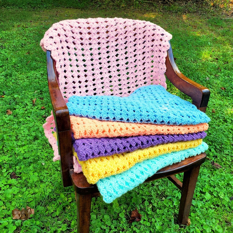 Pu&ograve; includere: Un mucchio di sei coperte all'uncinetto in vari colori pastello, tra cui rosa, blu, giallo e viola, sono esposte su una sedia di legno.