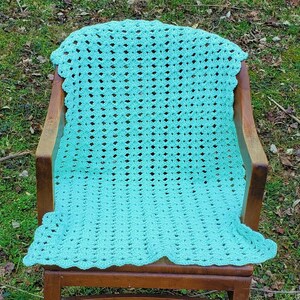 Pu&ograve; includere: Una coperta all'uncinetto color turchese con un bordo a festoni, drappeggiata su una sedia di legno.