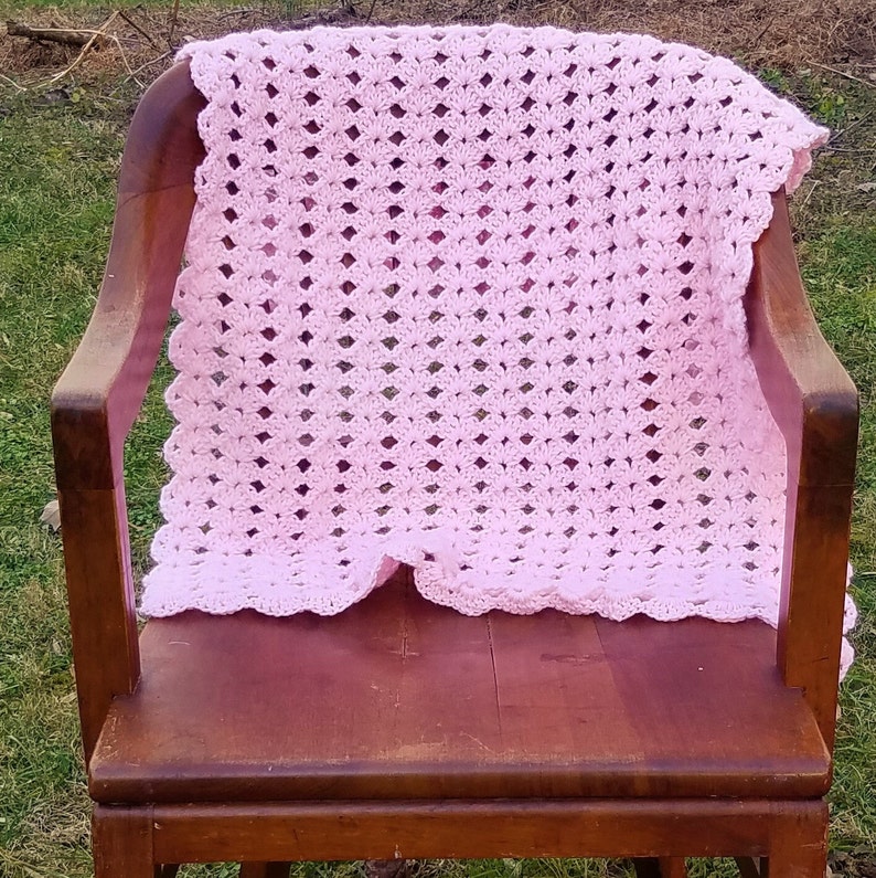 Pu&ograve; includere: Una copertina per neonati in crochet rosa con un bordo a festoni, drappeggiata su una sedia di legno.