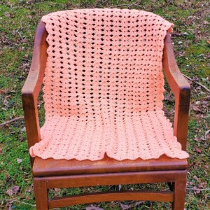 Pu&ograve; includere: Una coperta di uncinetto color pesca drappeggiata su una sedia di legno. La coperta ha un delicato motivo a pizzo ed &egrave; realizzata con filato morbido.