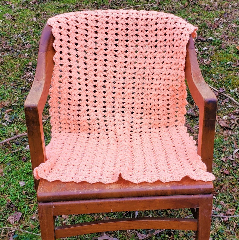 Pu&ograve; includere: Una coperta di uncinetto color pesca drappeggiata su una sedia di legno. La coperta ha un delicato motivo a pizzo ed &egrave; realizzata con filato morbido.